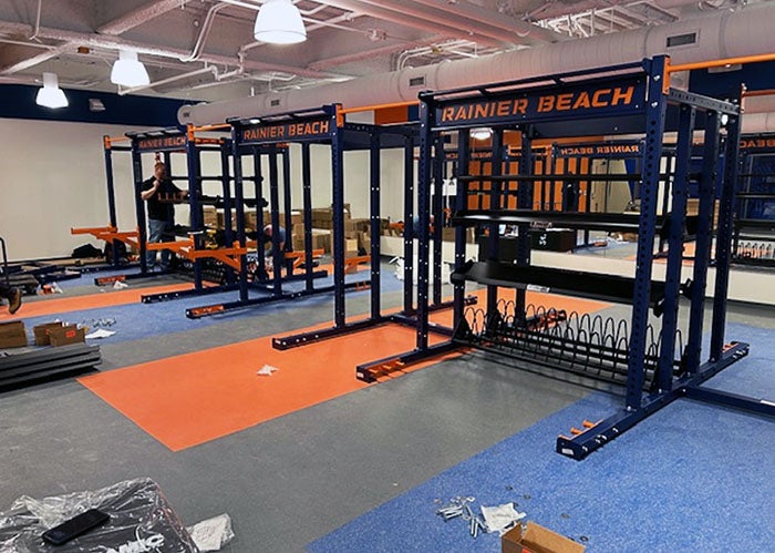 Fitness room with custom “Rainier Beach” branded weight racks installed on blue and orange flooring.